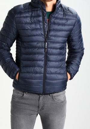 Mann trägt eine marineblaue gesteppte Jacke mit Frontreißverschluss und Händen in den Taschen, kombiniert mit hellgrauen Jeans.