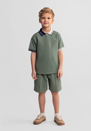 Młody chłopiec stoi, ubrany w zieloną krótką koszulkę polo z granatowo-białym kołnierzykiem, dopasowane zielone szorty, białe skarpetki i beżowe tenisówki.