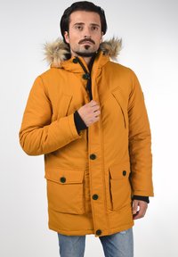 Narančasta izolirana parkas jakna s kapuljačom od krzna, zelenim gumbima, dva prednja džepa i zatvaračem. Tekstura tkanine i topli dizajn.
