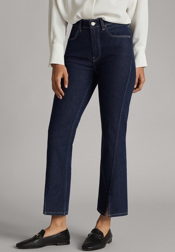 Femme portant un jean taille haute bleu marine avec des détails de coutures sur le devant, de légères fentes aux chevilles, un chemisier blanc et des mocassins noirs.