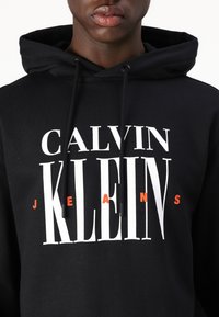 Juodas hoodis, kuriame yra didelis baltas ir oranžinis "Calvin Klein Jeans" logotipas. Medžiaga atrodo švelni, su raišteliu reguliuojamu gobtuvu.