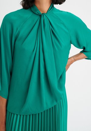 Blouse bleu-vert avec un col haut et un devant drapé, fabriquée dans un tissu doux, avec des manches trois-quarts et une texture lisse.
