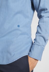 Ljusblå skjorta med knäppning som har en subtil struktur, med en liten logotyp på vänster sida och detaljer vid ärmsluten. Designad med en strömlinjeformad passform.