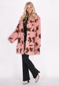 Abrigo de pelo sintético rosa con un patrón de manchas marrones, que cuenta con un cierre de botón y mangas anchas. Combinado con pantalones negros acampanados y tacones.