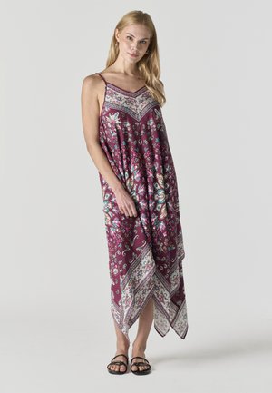 Mujer con cabello largo y rubio, vestida con un vestido maxi sin mangas color granate con patrones florales y geométricos, sandalias negras, de pie sobre un fondo claro.