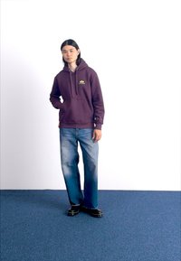 Bordo mikina s predným vreckom a žltým logom, v kombinácii s modrými roztrhnutými širokými džínami a čiernymi topánkami, na pozadí s bielym plátom.