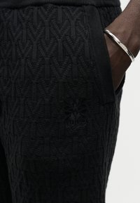 Κοντινό ενός χεριού στην τσέπη μαύρου υφασμάτινου παντελονιού με ασημένιο βραχιόλι εγχάρακτο με τη λέξη "HUGO".