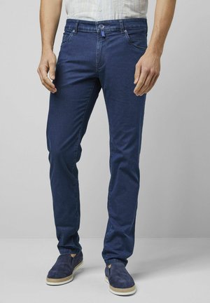 Man die een donkerblauwe slim-fit jeans en marineblauwe instapschoenen draagt tegen een effen lichtgrijze achtergrond.