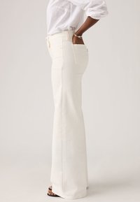 Άτομο που φοράει ψηλόμεσα, φαρδιά λευκά παντελόνια με λευκό πουκάμισο, χέρι στην πίσω τσέπη και μαύρα σανδάλια με ανοιχτά δάχτυλα.