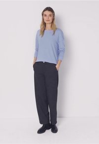 Nainen seisoo käsineen taskuissa, päällään vaaleansininen pitkähihaisen paita, tummat leveälahkeiset housut ja mustat nilkkurit valkoista taustaa vasten.