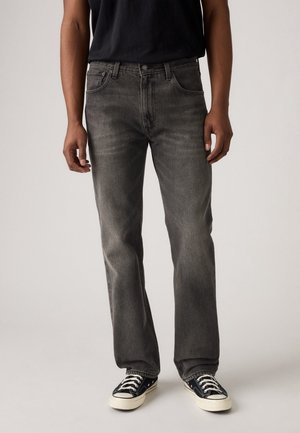 Personne portant un jean noir délavé à jambe droite, des baskets noires et blanches à lacets et une chemise noire, debout devant un fond uni.