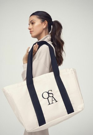 Žena s tmavým copom oblečená v béžovom oblečení, nesie veľkú bielu tašku s čiernymi popruhmi a vyšívaným logom "OSLA".