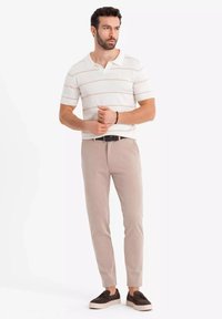 Valkoinen polo-paita vaaleilla raitoja, beige slim-fit-housut ja tummat kengät, seisoo kädet hieman ristissä neutraalia taustaa vasten.