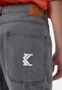 Harmaan denim-housut, joissa on nahkaläppä vyötäröllä ja valkoinen kirjailtu logo takataskussa. Ominaisuuksina perinteinen ommel ja varusteet.