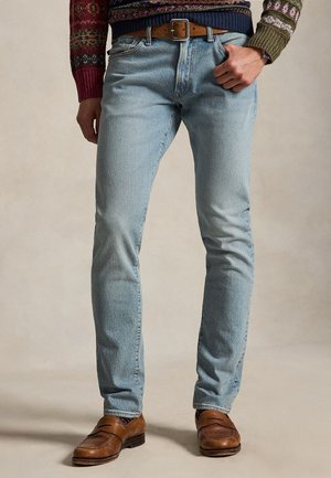 Bărbat îmbrăcat în blugi bleu, curea din piele maro, pulover cu modele colorate și pantofi din piele maro, stând cu o mână în buzunar.