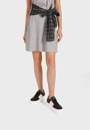 Donna che indossa un vestito grigio con una camicia a quadri bianca e nera annodata in vita, sneaker platform bianche e nere, in piedi su sfondo bianco.