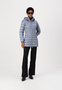 Giacca puffy grigio chiaro con chiusura a zip, cappuccio e cuciture orizzontali. Abbinata a pantaloni neri e stivali con tacco.