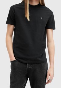 T-shirt nera a maniche corte in tessuto di cotone con un piccolo logo grigio sul petto. Abbinata a jeans grigio scuro.