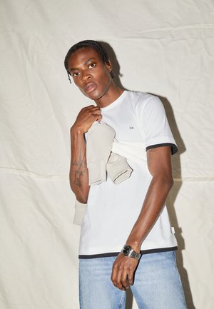T-shirt bianca a maniche corte con scollatura nera e logo 'CK', abbinata a un maglione arrotolato color beige chiaro. Modello che indossa jeans e un orologio d'argento.