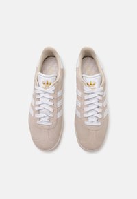 Baskets en suède beige avec des rayures blanches, dotées de lacets blancs et d'une semelle intérieure texturée, accentuées par un logo doré sur la languette.