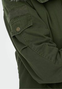 Manche d'une veste verte avec une poche boutonnée et un détail de sangle en Velcro, montrant le tissu plié et les motifs de couture.