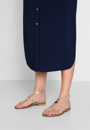 Fødder iført gyldne T-rem flade sandaler sammen med en navyblå midinederdel, stående på en glat grå gulv.
