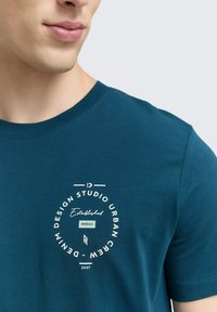 Žydros spalvos medvilnės marškinėliai, kuriuose yra balta apvali grafika su užrašu "Design Studio Urban Crew" ir rodyklės dizainu. Trumpomis rankovėmis ir apvaliu kaklu.