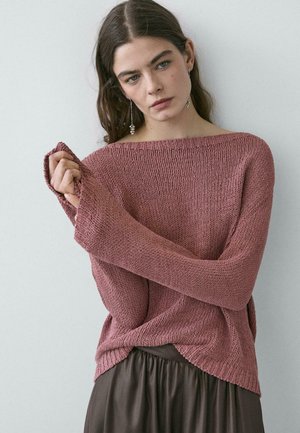 Giovane donna con lunghi capelli castani che indossa un maglione lavorato a maglia color malva e una gonna scura, in piedi contro una parete chiara e semplice.