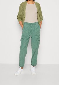 Lichtgroene cargobroek met twee zijzakken, taps toelopende enkels en een geweven textuur, gecombineerd met een beige top en witte sneakers.