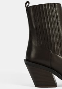 Bottine en cuir marron foncé avec des coutures verticales, un talon légèrement évasé et une finition brillante. Elle possède une languette à l'arrière.