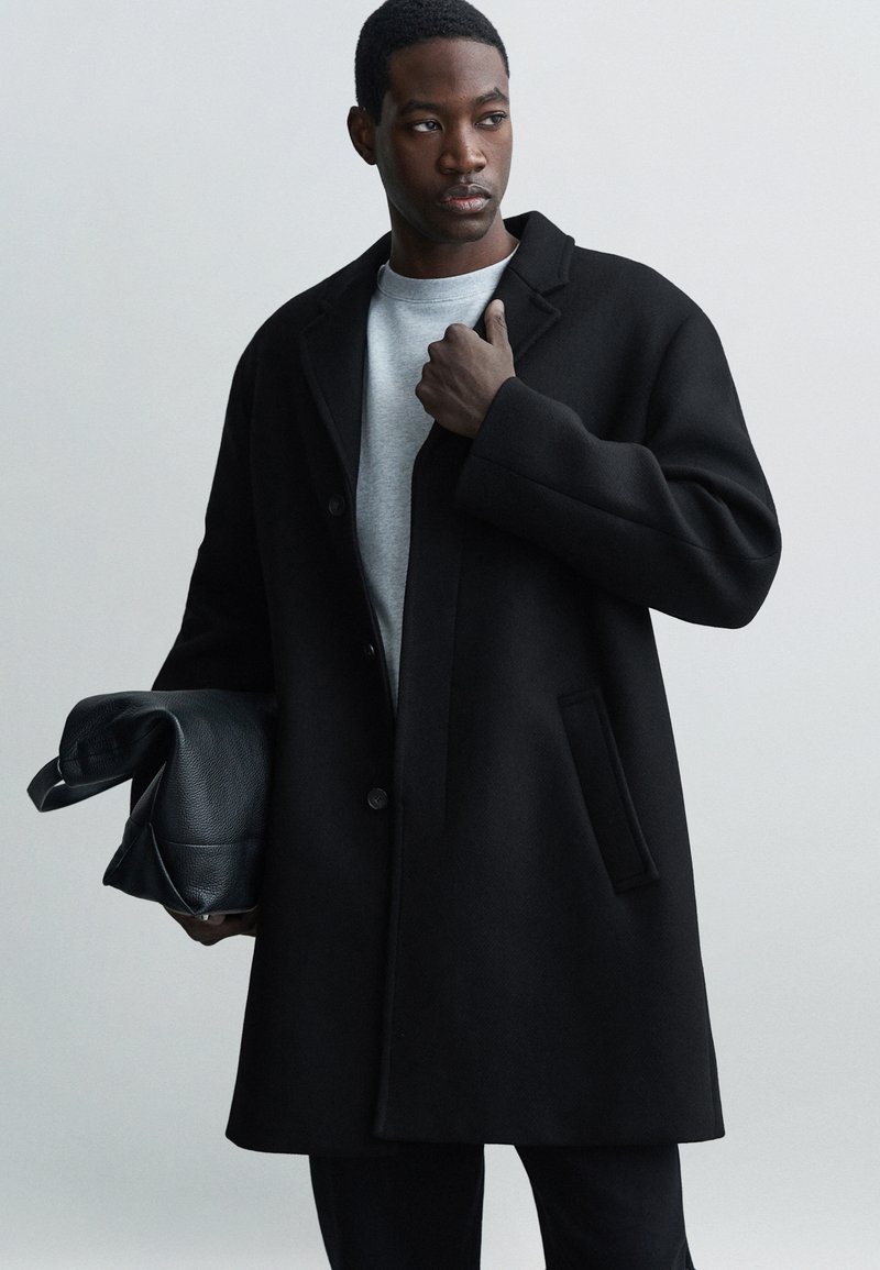 Fekete gyapjúpulóver, strukturált dizájnnal, nagy gallérral, elülső gombokkal és oldalsó zsebbel. Szürke pulóver fölött viselve, fekete táskával párosítva.