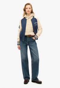 Marineblauwe en crèmekleurige varsityjas over een beige hoodie, gecombineerd met high-waisted blauwe jeans en donkere loafers. Voorzien van strepen en logo's.