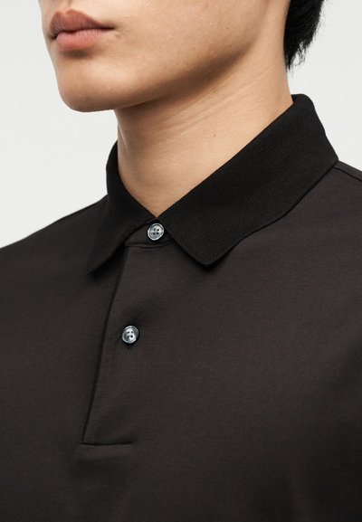 Εγγύς λήψη ενός ατόμου που φορά μαύρο πόλο μπλουζάκι με δύο ορατά κουμπιά και ριμπ γιακά πάνω σε απλό φόντο.