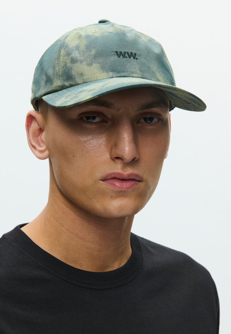 Camouflage baseball cap in green and gray tones, featuring a logo in black. The cap has a soft texture and a curved brim.
