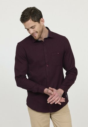 Chemise rouge bordeaux à manches longues avec un col à boutons, une poche poitrine unique et des accents de boutons contrastants. Le tissu semble doux avec une texture lisse.