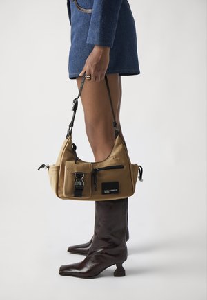 Tan nylon shoulder bag with black hardware, zip compartments, and adjustable straps, held by a person wearing a blue jacket and brown knee-high boots.
