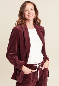 Burgunderfarbener Cord-Blazer mit entspannter Passform und offenem Vorderteil, kombiniert mit passenden Hosen, die einen Kordelzug und Vordertaschen haben.