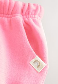 Tissu rose doux avec une ceinture élastique froncée et une petite étiquette blanche rectangulaire montrant un symbole gris incurvé.