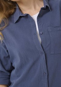 Mujer vistiendo una camisa azul desabrochada sobre una blusa blanca, con cabello rubio suelto visible en el lado izquierdo.