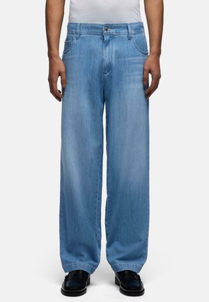 Uomo che indossa jeans azzurri a gamba larga e scarpe nere, in piedi davanti a uno sfondo bianco uniforme.