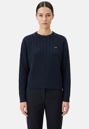 Donna con i capelli scuri raccolti indietro, che indossa un maglione a trecce blu navy e pantaloni neri, in piedi davanti a uno sfondo bianco uniforme.