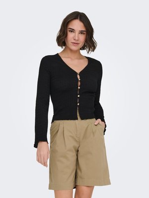 Femme aux cheveux bruns courts portant un cardigan noir ajusté à boutons et un short plissé beige mi-cuisse, les mains dans les poches.