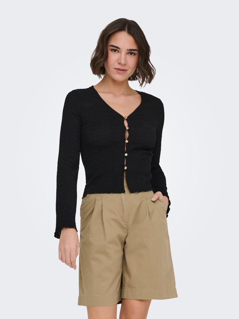 Donna con capelli castani corti che indossa un cardigan nero aderente con bottoni e pantaloncini beige plissettati fino al ginocchio, con le mani nelle tasche.