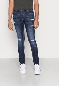 Vaqueros ajustados de color azul oscuro con áreas desgastadas y detalles envejecidos, con salpicaduras de pintura blanca. Combinados con zapatillas blancas.