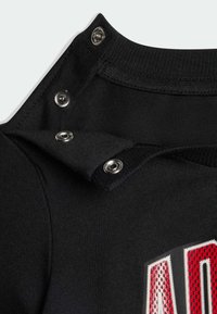 Cuello de camiseta negra con botones a presión plateados; la textura de la tela es suave. Cuenta con un diseño gráfico rojo con elementos texturizados.