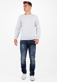 Sweatshirt gris à col rond avec des manches longues, associé à un jean en denim bleu foncé. Chaussures blanches, se tenant sur un fond blanc.