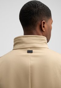 Tan hoge kraag jas met een soepele textuur, voorzien van een zwart label op de achterhals en een nette stiksels op de naden.