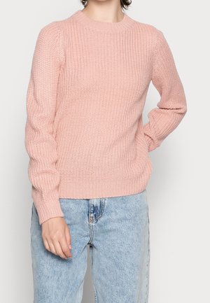 Person iført en lyserød strikket sweater med lange ærmer og lyseblå jeans, der poser med den ene hånd på hoften.