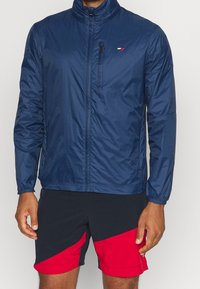 Chaqueta ligera marina con cierre delantero, cuello alto y puños elásticos. Presenta un pequeño logo y un bolsillo en el pecho con cremallera.