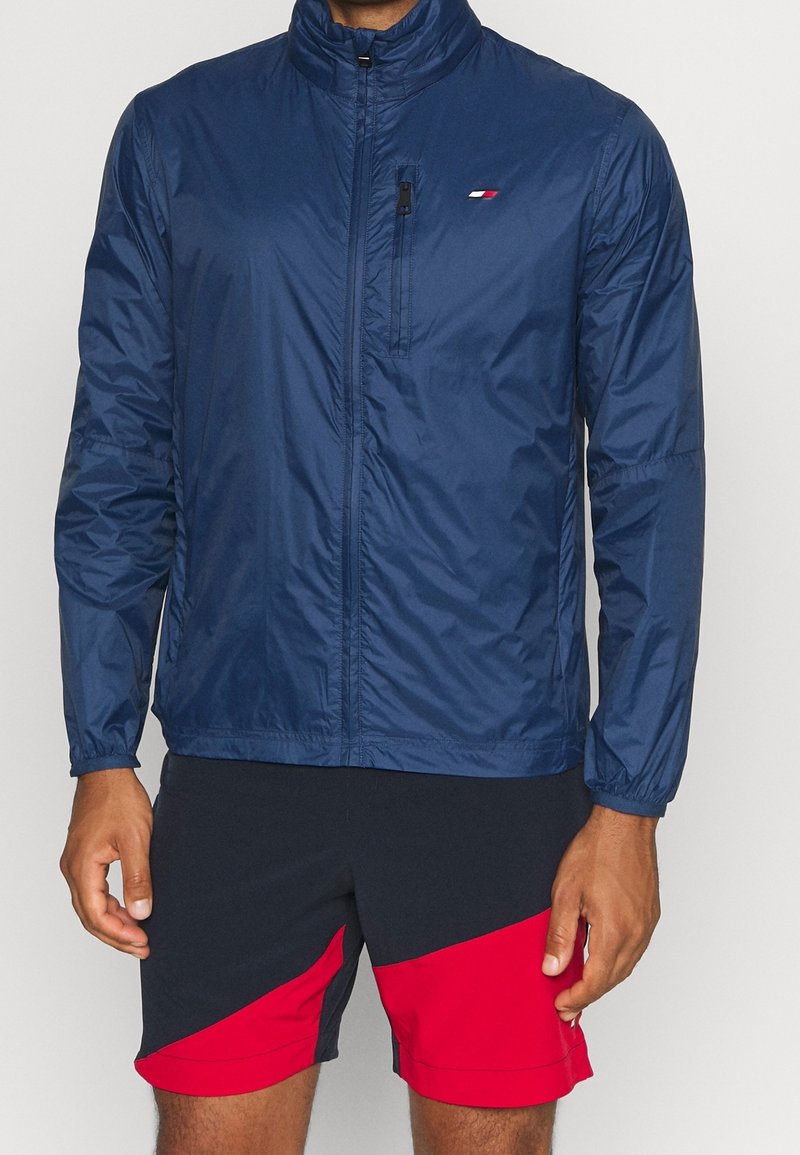Chaqueta ligera marina con cierre delantero, cuello alto y puños elásticos. Presenta un pequeño logo y un bolsillo en el pecho con cremallera.